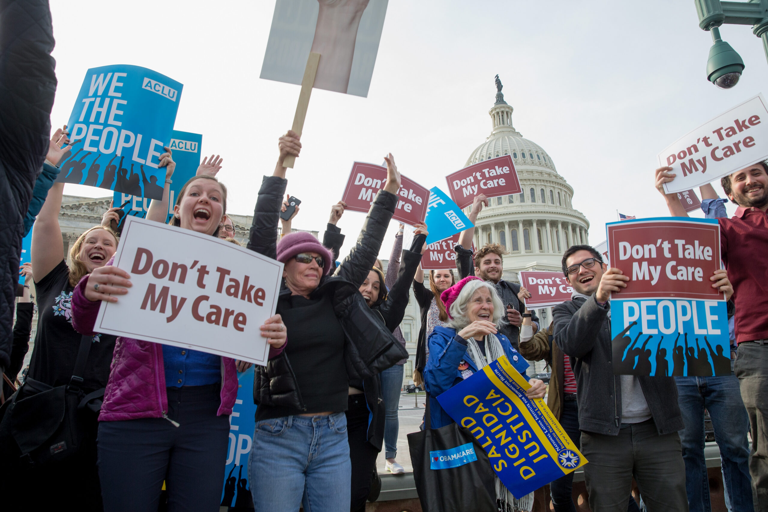 A group of people smiling and holding up signs against cuts to healthcare funding while at a rally in front of the United States Capitol building.