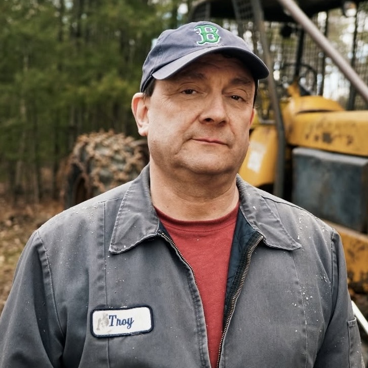 This is an image of Troy Jackson, candidate for Maine Senate. He is pictured in a grey jacket with a red t-shirt standing in front of a tractor in a forest.