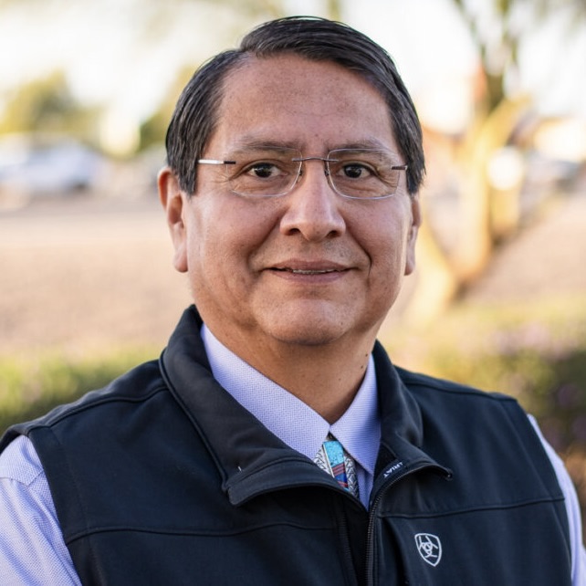 This is an image of Jonathan Nez, candidate for Arizona's 2nd Congressional District. He is pictured in a black vest with a purple shirt in front of some greenery.